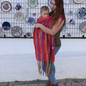 Mujer porteando en una bandolera indajani yee rojo a un bebé grande con tonos cálidos con fondo platos de cerámica