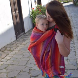 Mujer porteando en una bandolera indajani yee rojo a un bebé grande con tonos cálidos con fondo de calle antigua