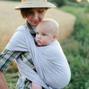 Madre con un sombrero porteando a su bebé en un fular little frog lovely heath de fondo un prado.