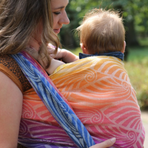 fotografía de lado de una Mujer porteando a un bebé con el fular tejido Yaro Dandy Havana Grad, en un parque mostrando el degradado de colores desde el morado al naranja.