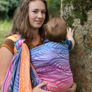 Mujer porteando a un bebé con el fular tejido Yaro Dandy Havana Grad, en un parque mostrando el degradado de colores desde el morado al naranja.