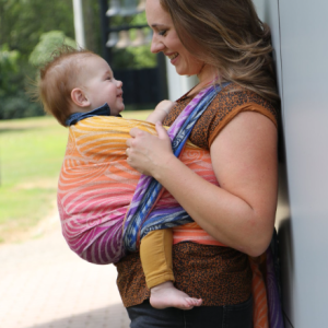 Mujer porteando a un bebé de lado con el fular tejido Yaro Dandy Havana Grad, mostrando el degradado de colores desde el morado al naranja en un parque.