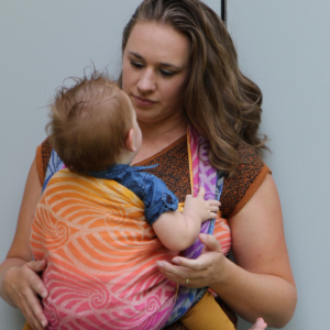 Mujer porteando a un bebé con el fular tejido Yaro Dandy Havana Grad, mostrando el degradado de colores desde el morado al naranja en un parque.