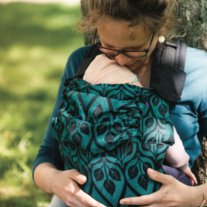 Madre besando tiernamente a su bebé porteado en una mochila ergonómica Yaro Flex, con el diseño de hojas 'La Vita' en colores negro y verde esmeralda