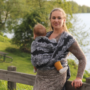 Madre porteando a su bebé al aire libre con el fular tejido Yaro Jazz Duo Black, con un diseño de flores en blanco y negro. apoyada en una vaya de madera