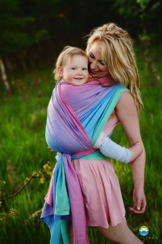Madre riendo porteando a su hija en un campo verde con el fular Little Frog Lolite, de tejido mezcla con Tencel en colores arcoíris pastel.