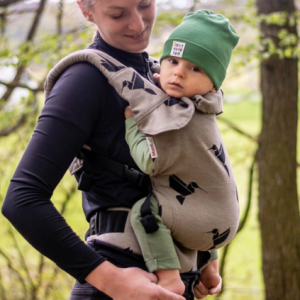 Madre porteando a su bebé en un bosque con la mochila ergonómica MoniLu Uni Start, de color arena con estampado de colibríes negros