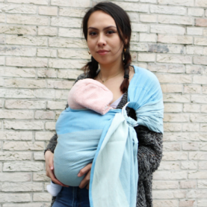 Madre porteando a su bebé recién nacido (con gorrito rosa) en una bandolera de anillas Yaro Blue (azul cielo), mostrando un ajuste alto y seguro en el pecho.