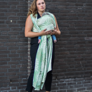 Madre de pie frente a una pared de ladrillo oscuro usando un fular de porteo verde claro, mostrando el soporte correcto en la espalda y hombros.