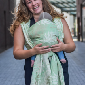 Madre paseando sonriente con su bebé en el pecho, envueltos en el fular Oasis Duo que combina hilos verdes y crudos para un efecto luminoso.