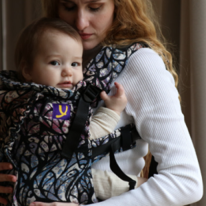 Madre porteando a su bebé de 2 meses en una mochila ergonómica Yaro Hug, con diseño 'Reality of Dreams' de estilo mosaico colorido sobre fondo negro.