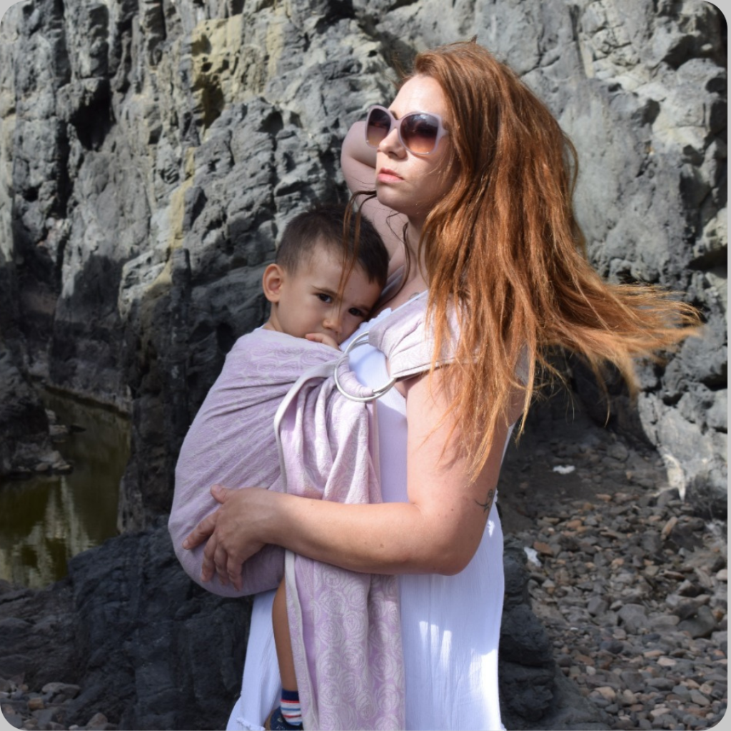 Madre porteando a la cadera a su hijo pequeño en una bandolera de anillas de tejido de fular rosa sobre un fondo de rocas al aire libre.