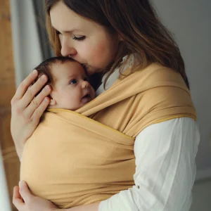 Plano medio de madre besando la cabeza de su recién nacido acurrucado ergonómicamente en fular semielástico mostaza Little Frog Honey Sun.