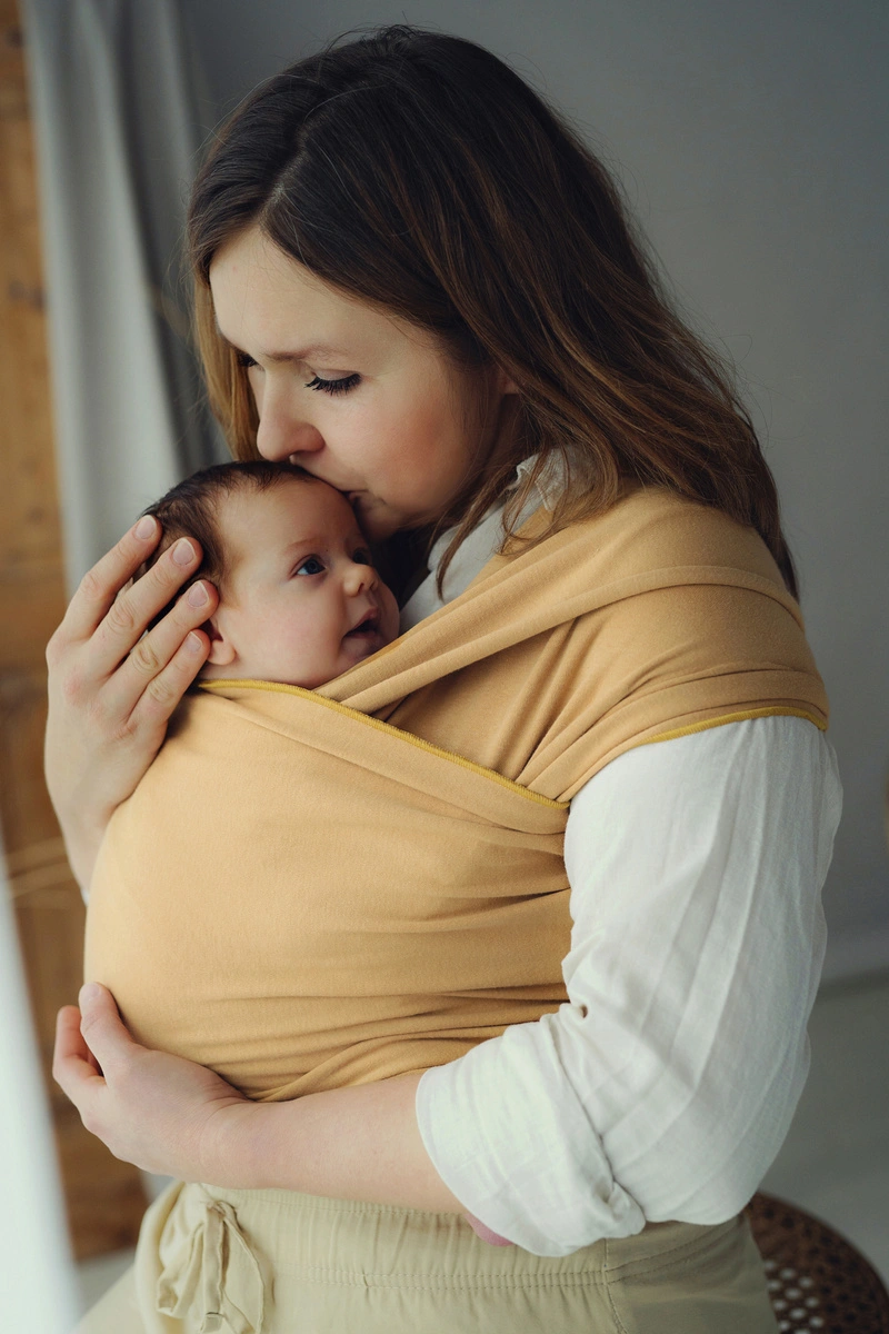 Plano medio de madre besando la cabeza de su recién nacido acurrucado ergonómicamente en fular semielástico mostaza Little Frog Honey Sun.