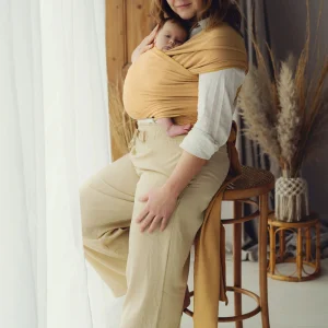 Madre de pie posando junto a un ventanal con cortina blanca portando a su bebé recién nacido en fular semielástico amarillo mostaza.