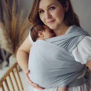 Madre sonriendo a cámara sentada con su bebé recién nacido dormido sobre su pecho en fular semielástico Little Frog color gris claro Misty Grey.