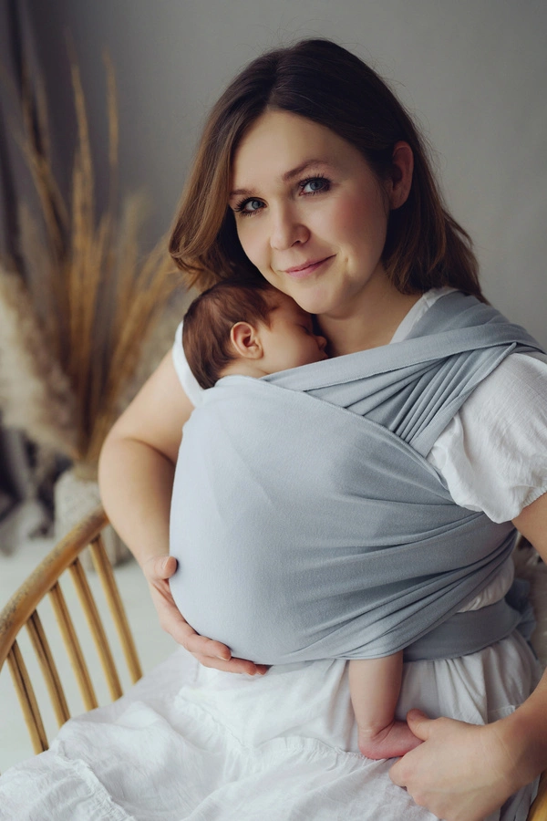 Madre sonriendo a cámara sentada con su bebé recién nacido dormido sobre su pecho en fular semielástico Little Frog color gris claro Misty Grey.
