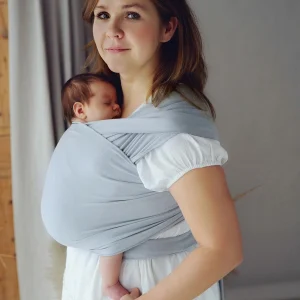Madre de pie mirando al frente mientras sostiene a su bebé recién nacido dormido en posición vertical ergonómica con fular semielástico gris.