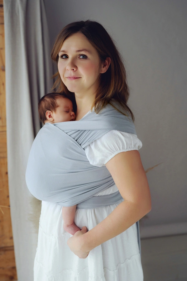 Madre de pie mirando al frente mientras sostiene a su bebé recién nacido dormido en posición vertical ergonómica con fular semielástico gris.
