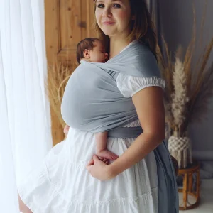 Madre vestida con vestido blanco posando de pie junto a un ventanal con su bebé en el fular semielástico gris Little Frog Misty Grey.