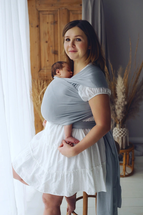 Madre vestida con vestido blanco posando de pie junto a un ventanal con su bebé en el fular semielástico gris Little Frog Misty Grey.