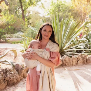 Madre con vestido rosa paseando por un jardín botánico porteando a su bebé en la bandolera de anillas Boba Ring Sling color Pampas Beige.