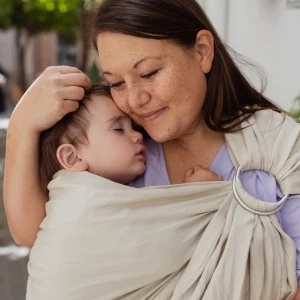 Plano corto de madre apoyando su cara en la cabeza de su bebé dormido plácidamente en la bandolera de anillas Boba Ring Sling Pampas Beige.