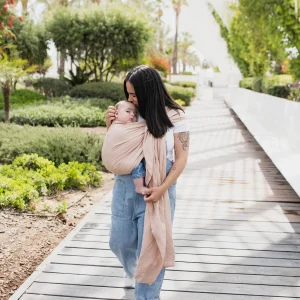 Madre caminando por una pasarela de madera rodeada de vegetación mientras portea a su bebé en la bandolera de anillas Boba Ring Sling en tono rosa empolvado Peony Pink.