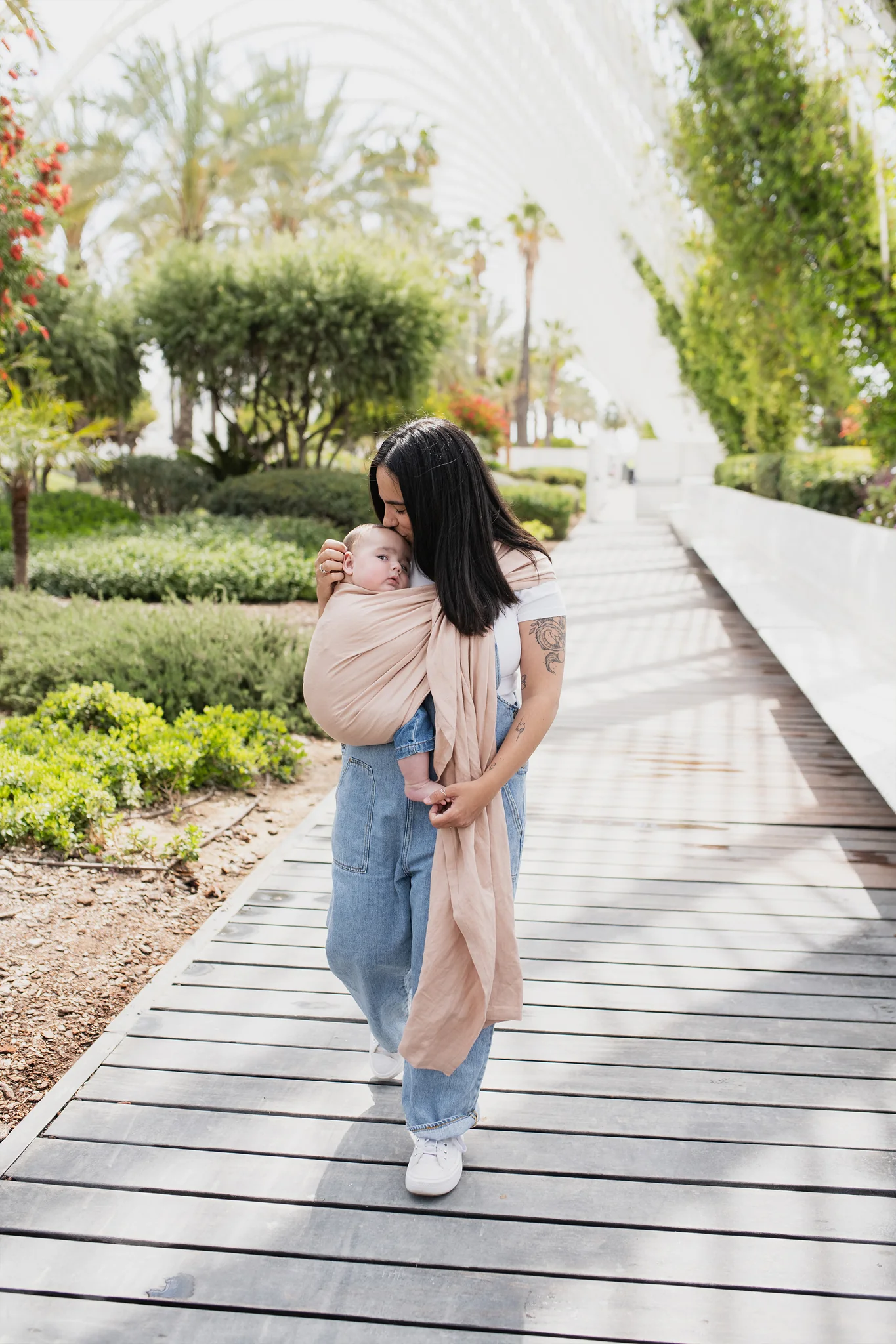 Madre caminando por una pasarela de madera rodeada de vegetación mientras portea a su bebé en la bandolera de anillas Boba Ring Sling en tono rosa empolvado Peony Pink.