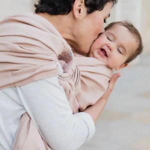 Plano medio de madre dando un beso lleno de ternura en la mejilla de su bebé sonriente porteado en la bandolera de anillas Boba Ring Sling Peony Pink.