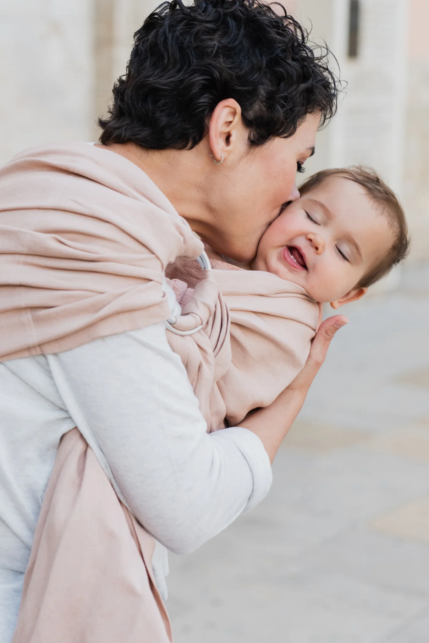 Plano medio de madre dando un beso lleno de ternura en la mejilla de su bebé sonriente porteado en la bandolera de anillas Boba Ring Sling Peony Pink.