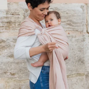 Madre con pelo corto ajustando la tela de la bandolera de anillas Boba Ring Sling Peony Pink mientras sostiene a su bebé junto a una pared de piedra.