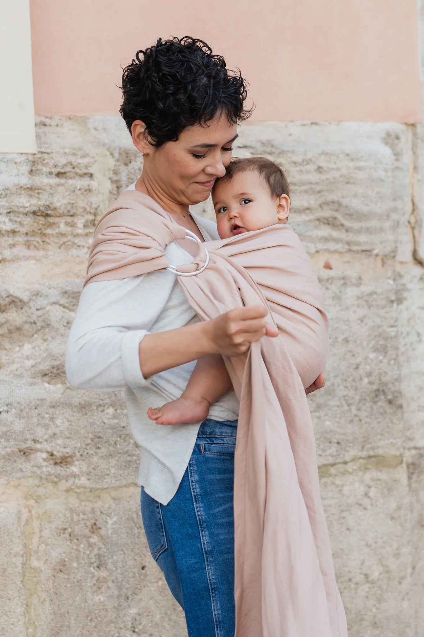 Madre con pelo corto ajustando la tela de la bandolera de anillas Boba Ring Sling Peony Pink mientras sostiene a su bebé junto a una pared de piedra.