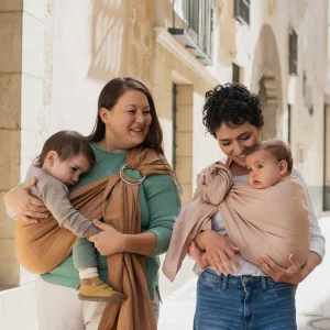 Dos madres caminando sonrientes por una calle empedrada porteando a sus bebés en bandoleras de anillas Boba Ring Sling en tonos marrón y rosa Peony Pink.