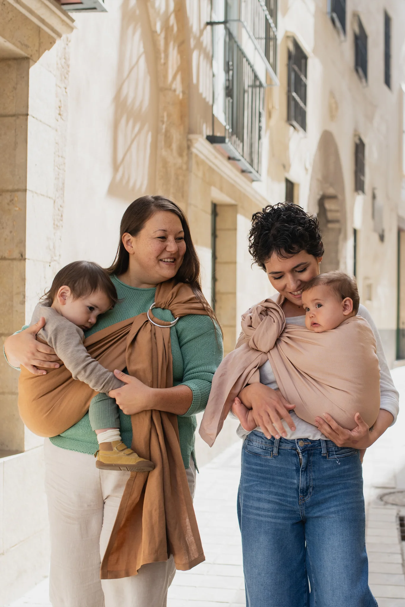 Dos madres caminando sonrientes por una calle empedrada porteando a sus bebés en bandoleras de anillas Boba Ring Sling en tonos marrón y rosa Peony Pink.