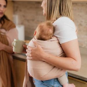 Plano medio de madre porteando a su bebé en la bandolera de anillas Boba Ring Sling Peony Pink en la cocina mientras sostiene una taza de café.