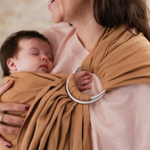 Plano detalle de madre sonriendo mientras sostiene a su recién nacido dormido en la bandolera de anillas Boba Ring Sling en color marrón Rye Brown, mostrando el ajuste en las anillas de aluminio.