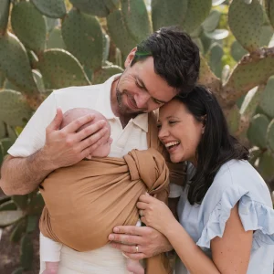 Padre porteando a su recién nacido en la bandolera de anillas Boba Ring Sling Rye Brown junto a la madre sonriendo al aire libre en un entorno natural.