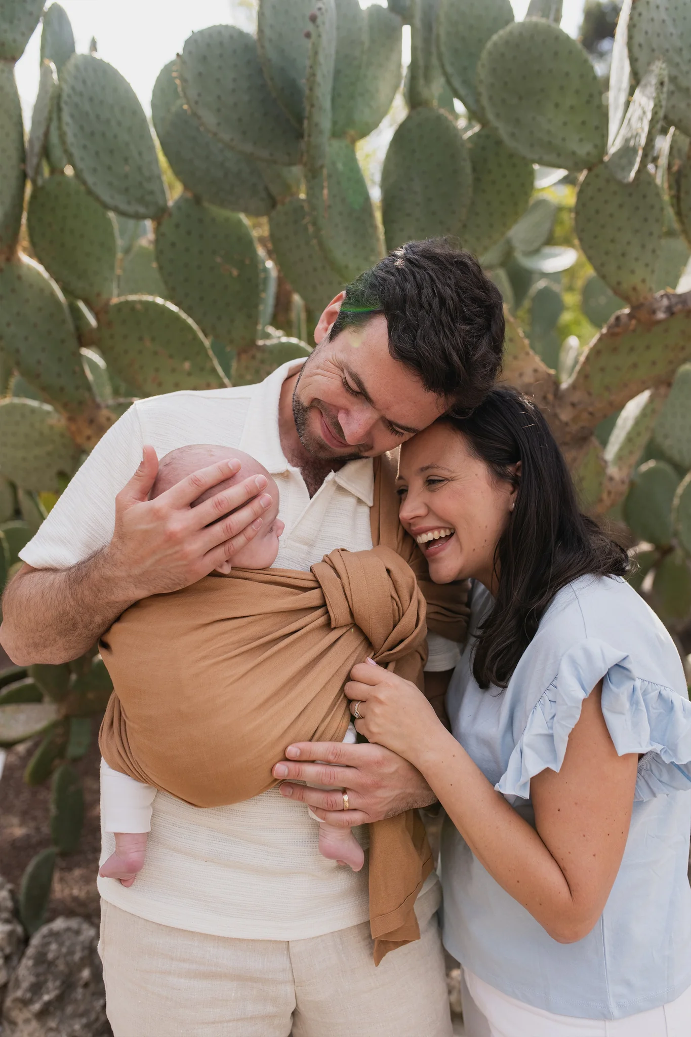 Padre porteando a su recién nacido en la bandolera de anillas Boba Ring Sling Rye Brown junto a la madre sonriendo al aire libre en un entorno natural.