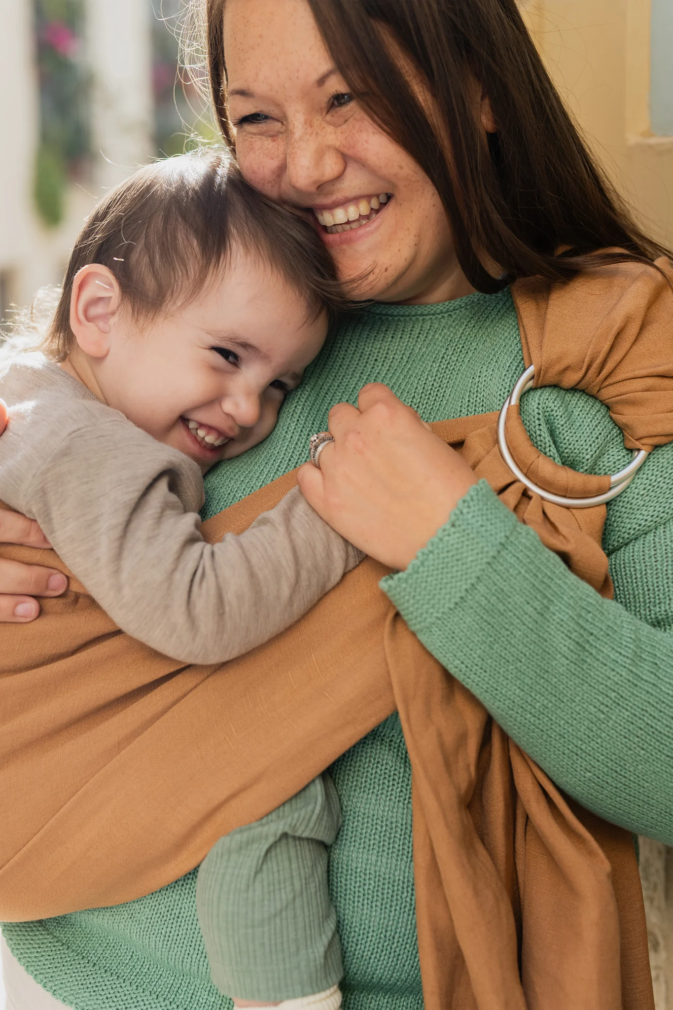 Madre e hijo mayor riendo juntos mientras el niño va porteado a la cadera en la bandolera de anillas Boba Ring Sling color marrón Rye Brown.