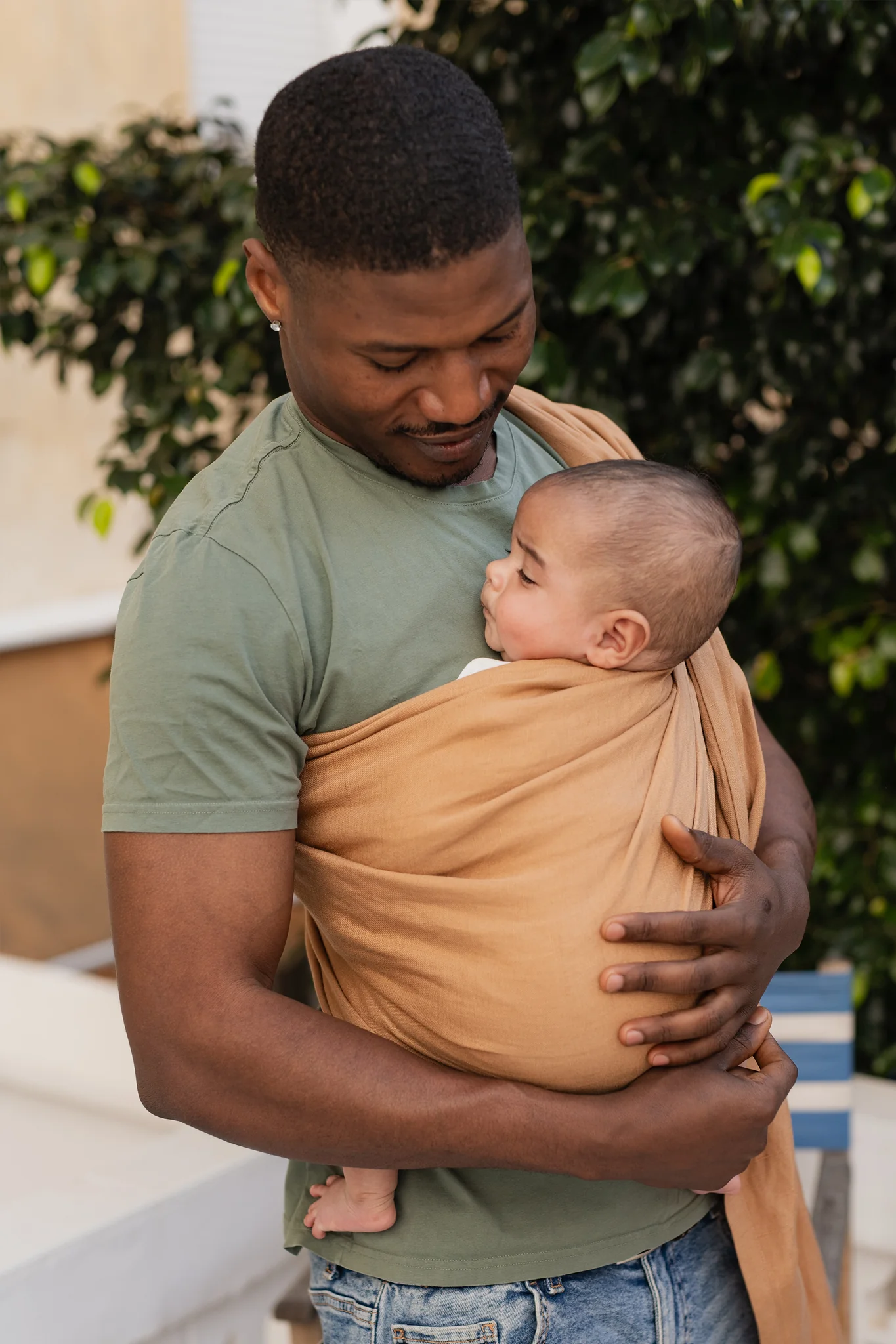Padre en camiseta verde mirando con amor a su bebé recién nacido dormido sobre su pecho en la bandolera de anillas Boba Ring Sling Rye Brown.