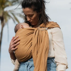Plano medio de madre de perfil al aire libre contemplando a su recién nacido alojado en la bandolera de anillas Boba Ring Sling tono marrón caramelo Rye Brown.