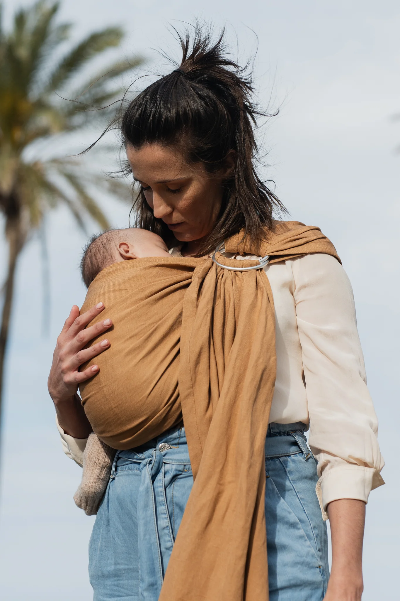 Plano medio de madre de perfil al aire libre contemplando a su recién nacido alojado en la bandolera de anillas Boba Ring Sling tono marrón caramelo Rye Brown.