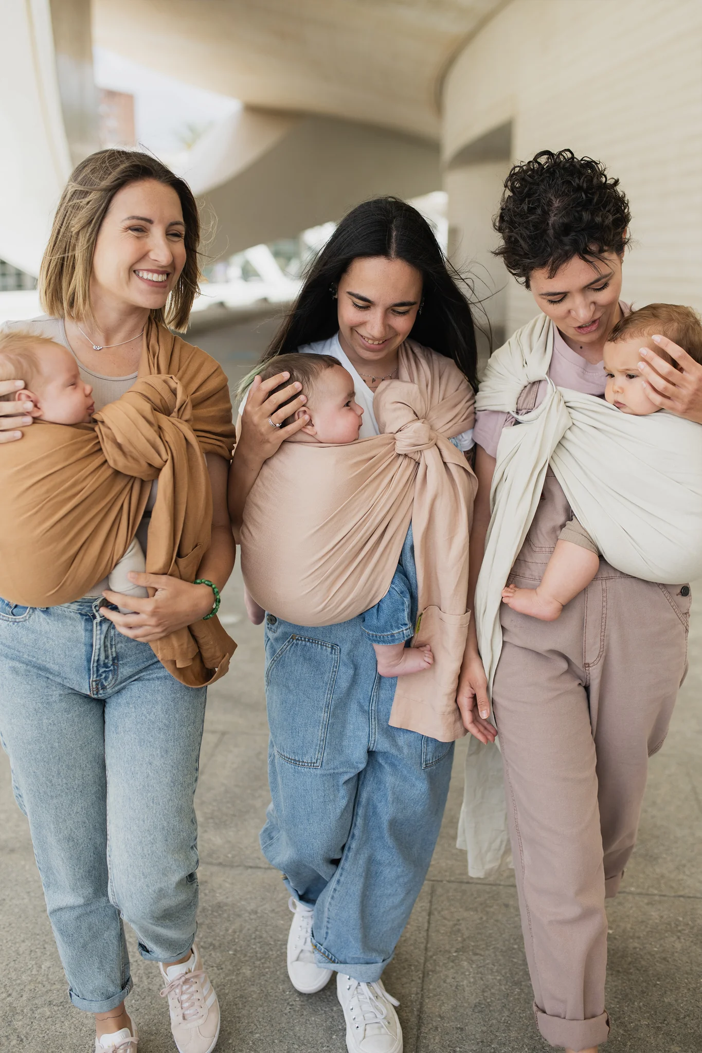 Tres madres sonrientes caminando juntas mientras portean a sus bebés con bandoleras de anillas Boba Ring Sling en tonos neutros Rye Brown y crema.