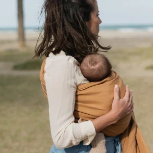 Vista trasera de madre caminando por la playa sujetando a su bebé porteado en la bandolera de anillas Boba Ring Sling Rye Brown.