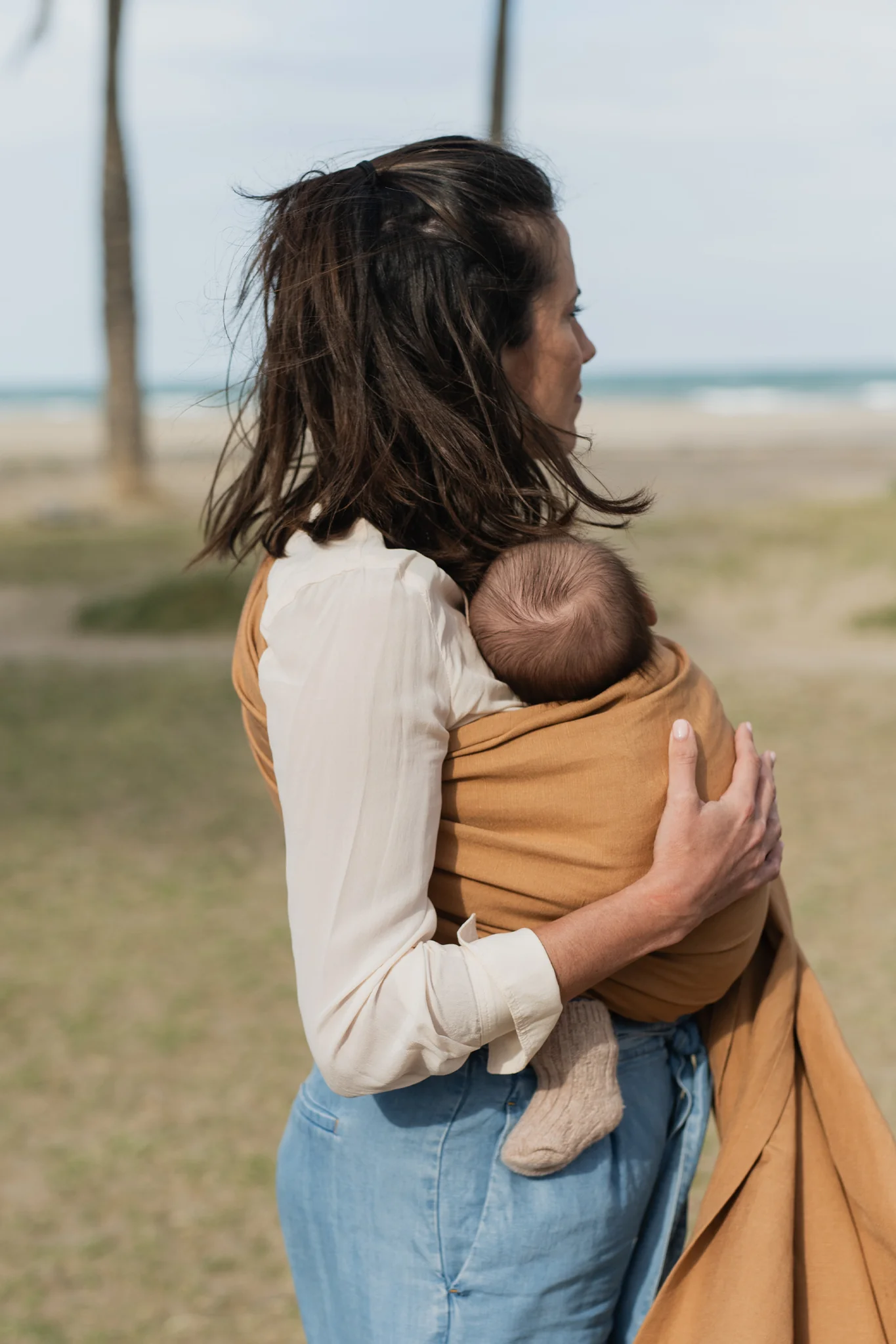 Vista trasera de madre caminando por la playa sujetando a su bebé porteado en la bandolera de anillas Boba Ring Sling Rye Brown.