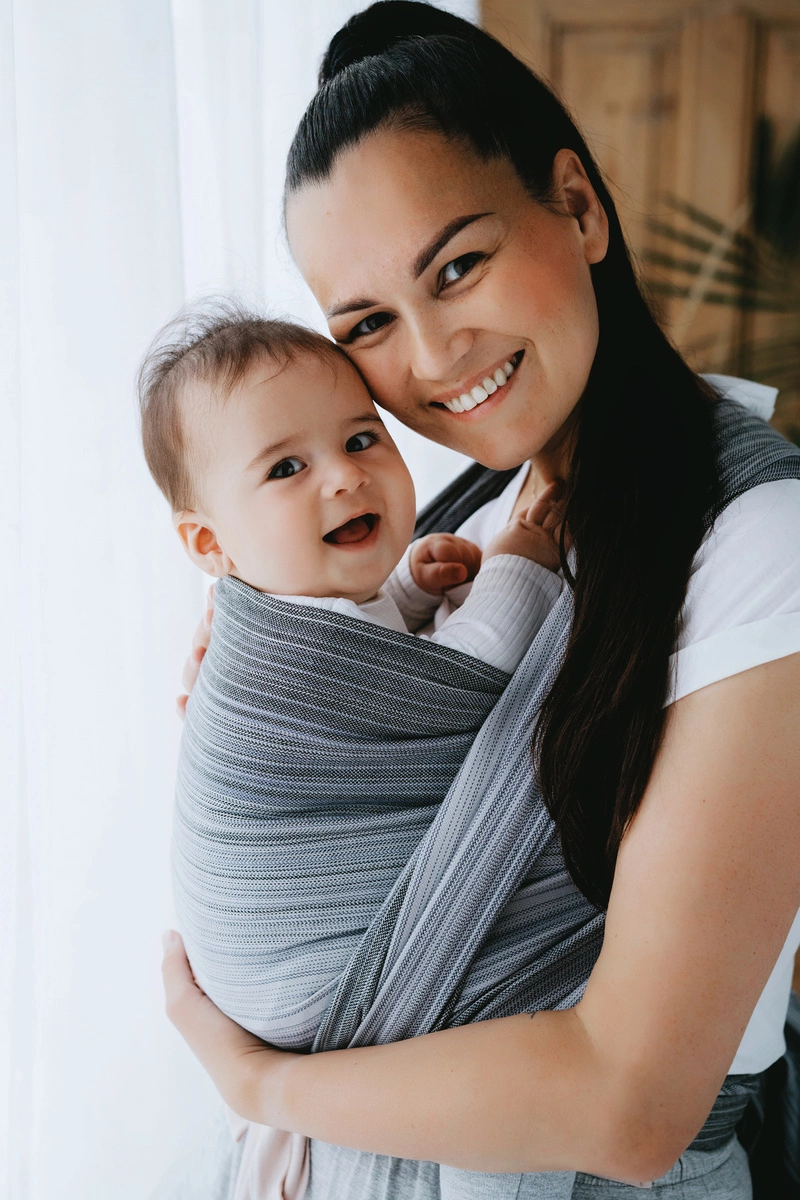 Plano corto de madre e hijo sonriendo alegremente a cámara mientras el bebé va sostenido ergonómicamente en el fular tejido gris Angelite.