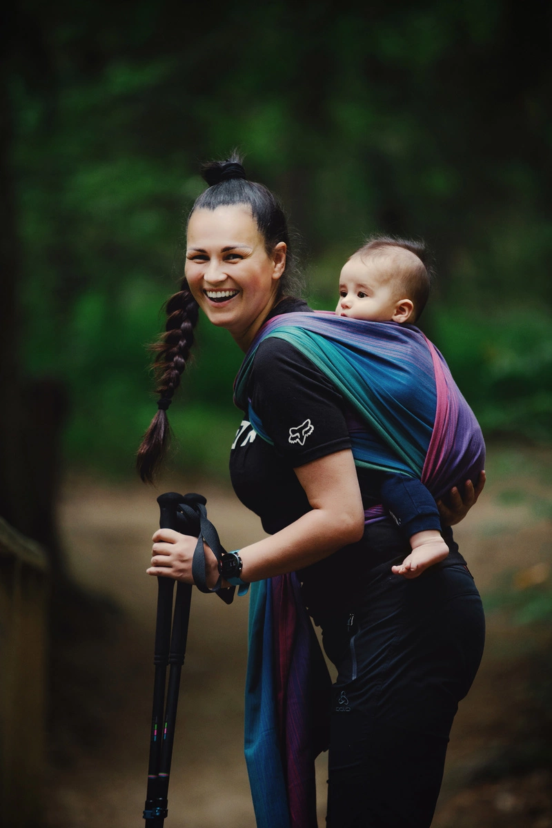 Plano medio de madre sonriendo alegremente a cámara sosteniendo bastones de trekking con su bebé ergonómicamente porteado en el fular Little Frog Misty Zircon.