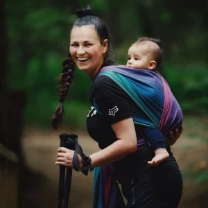 Plano medio de madre sonriendo alegremente a cámara sosteniendo bastones de trekking con su bebé ergonómicamente porteado en el fular Little Frog Misty Zircon.