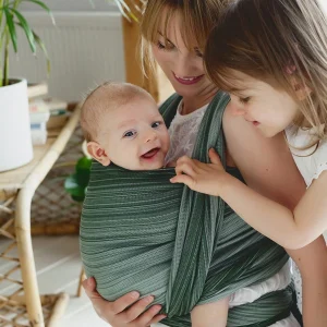 Madre e hija mayor sonriendo mientras la niña acaricia al recién nacido porteado al frente en el fular tejido de bambú Little Frog Verdelite en color verde sabio.